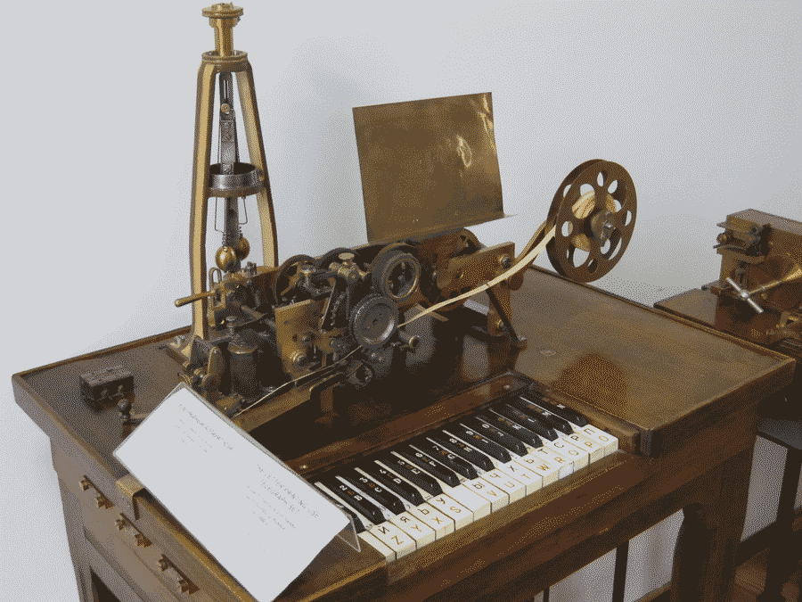 An image of an early telegraph machine, with piano keys acting as a keyboard. All components are uncovered, with big wheels and rollers sitting on top of the table which serves as a case for the system.