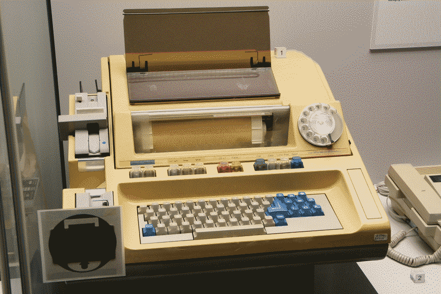 A late model teleprinter in a museum. It has rectangular shape and a wide keyboard, with additional components such as a telephone dial and a punch card reader on the sides.