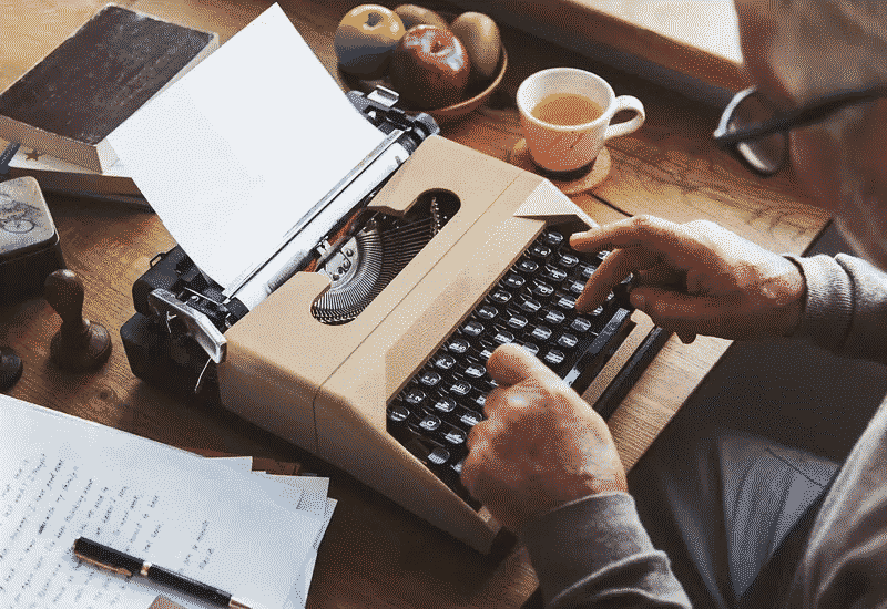 An old man sitting at a table in front of a minimalistic typewriter.
There is a bowl of fruit along with a small cup of tea on the table.
Several sheets of paper lay near the typewriter. Natural sunlight
shines down at the scene.